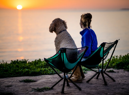 Woman and dog both sitting in cliq chairs watching the sun set over the ocean