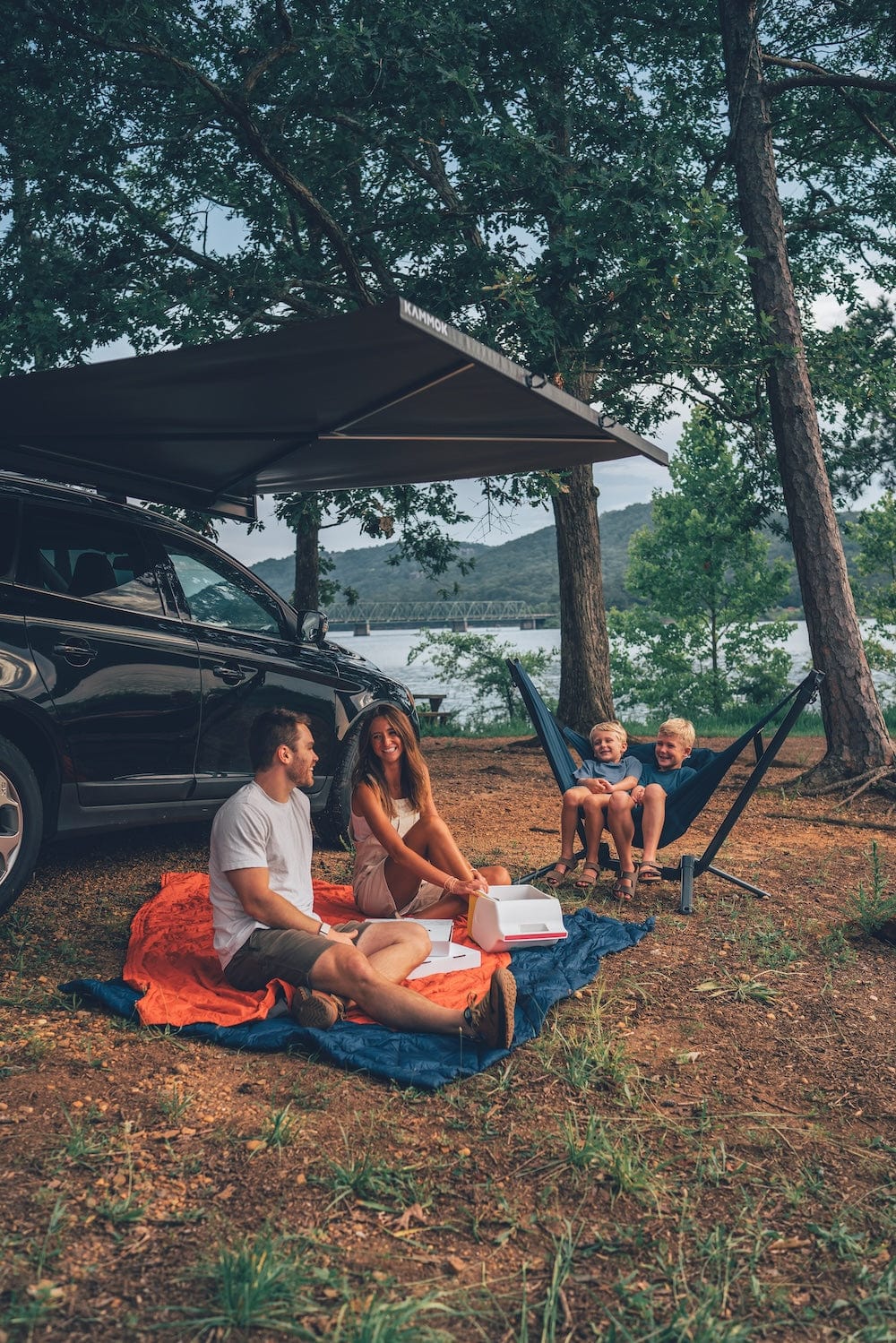 Family enjoying a picnic by a lake with a car and hammock stand and perch hammock chair in the background.