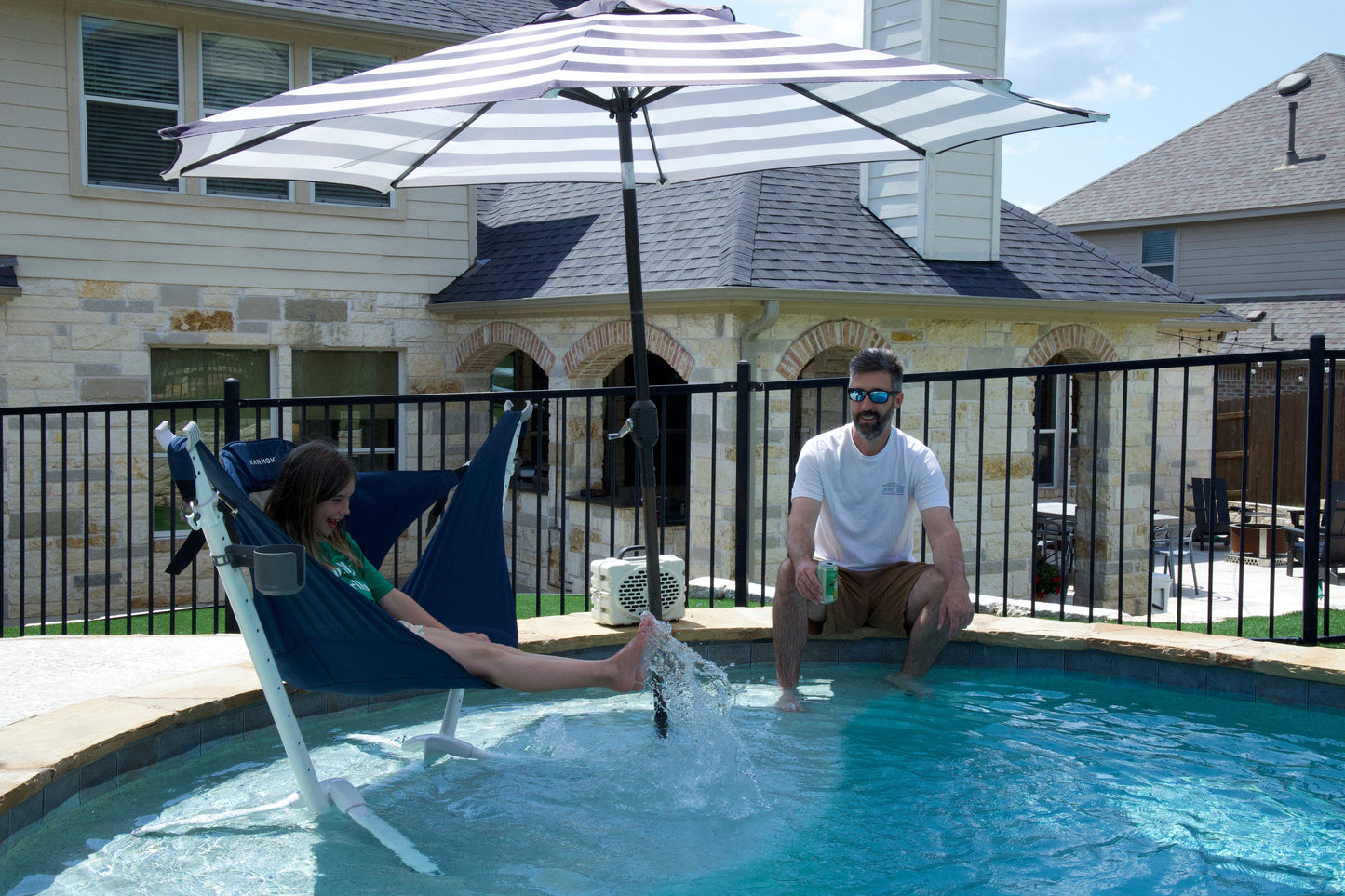 Two people by a pool with a striped umbrella with perch hammock chair and swiftlet hammock stand