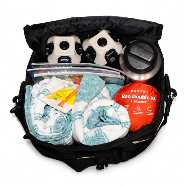 An open black backpack shown from above, filled with camping gear including two white YETI tumblers, a clear storage container, folded white towels with teal stripes, and an orange packaged Kammok Roo Double XL Hammock. - X-Large Chair Tote Bag