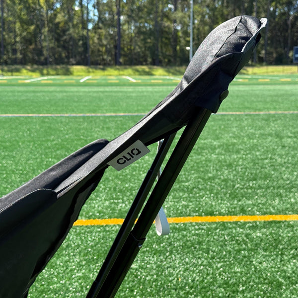 A close-up of a black outdoor lounge chair with a visible 'CLIQ brand tag, positioned on a green artificial turf field with trees in the background.
