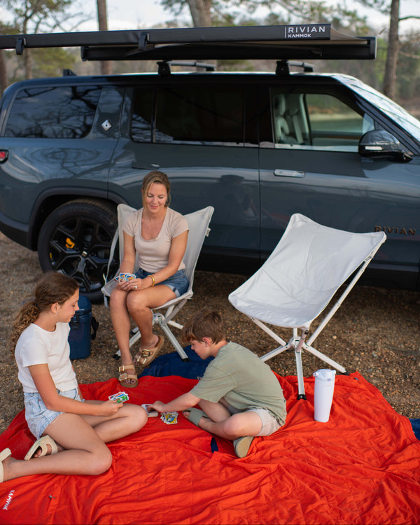 Family sitting on a red blanket in front of a black Rivian vehicle with a roof rack.