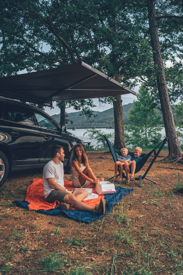 Family enjoying a picnic by a lake with a car and hammock stand and perch hammock chair in the background.