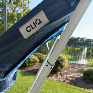 Close-up of a dark blue mesh canopy with a white metal frame and a visible CLIQ brand tag, set outdoors on grass with trees and a fence in the background. Limited Edition MystiQ Blue - CLIQ