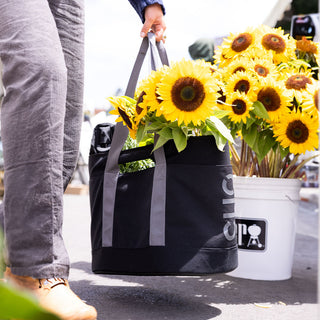 Person holding a black tote bag with sunflowers, standing next to a white bucket with sunflowers. - CLIQ Back Black