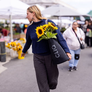 Woman carrying sunflowers and a black bag with CLIQ branding at an outdoor market.
