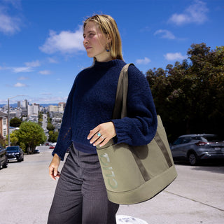 Woman in a blue sweater and gray pants holding a Olive tote ToQ bag on a city street.