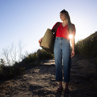 Woman walking outdoors holding a bag with a brand logo, sun shining behind her. Burnt Olive CLIQ Bag