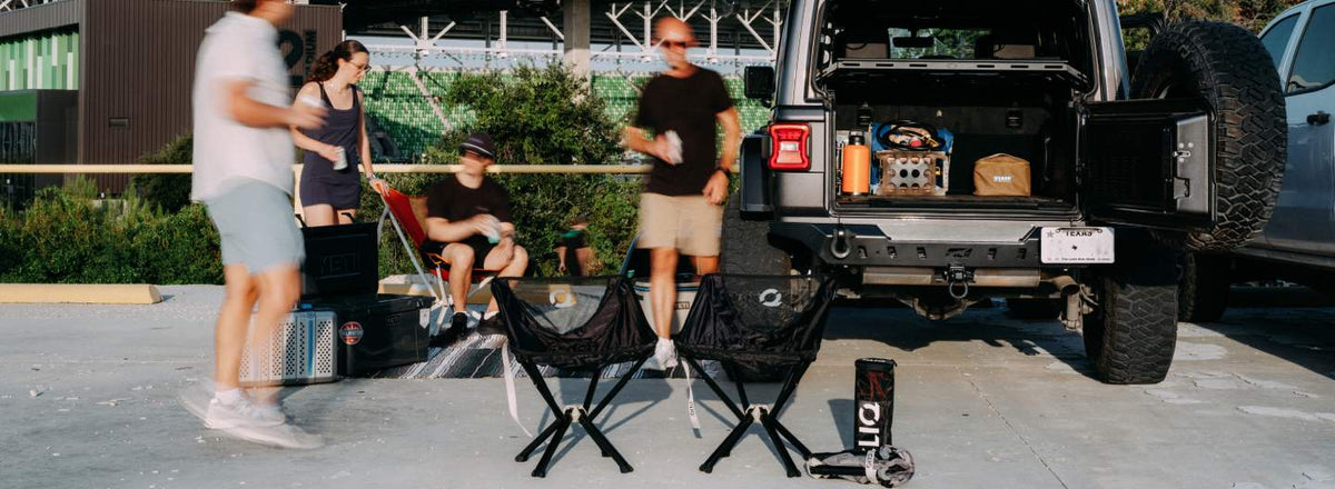 Outdoor lifestyle scene with several people standing and sitting near the open trunk of a black SUV. Two black folding CLIQ tailgatingchairs are placed in front, along with a cooler, bags, and a small black item standing on the ground.