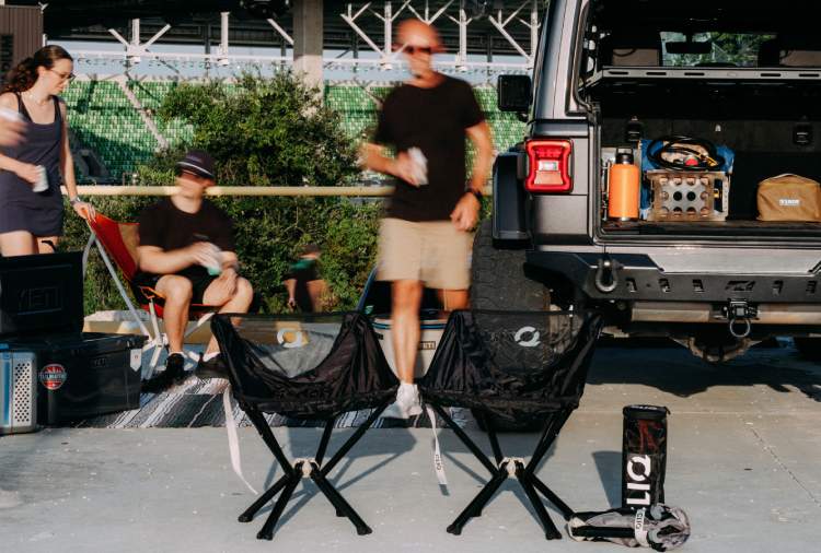 Two black folding CLIQ tailgating chairs in the foreground with a person standing nearby and other outdoor gear, including an open SUV trunk and tailgating items, visible in the background.